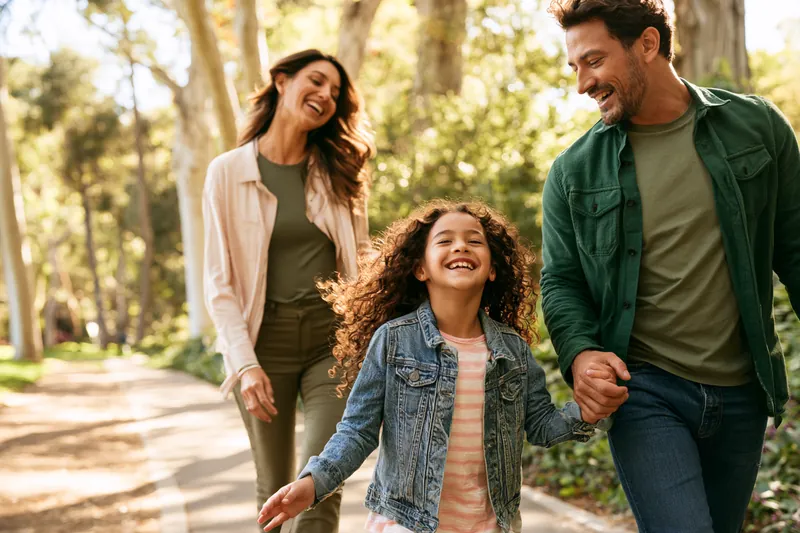Family of three walking hand in hand along a tree-lined park path, laughing together on a relaxed afternoon